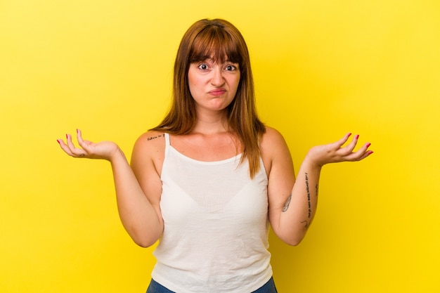 Giovane donna curvy caucasica isolata su sfondo giallo dubitando e alzando le spalle nel gesto interrogativo.