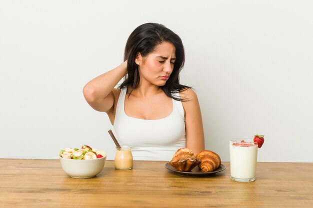 Giovane donna curvy che fa colazione che soffre di dolore al collo a causa dello stile di vita sedentario