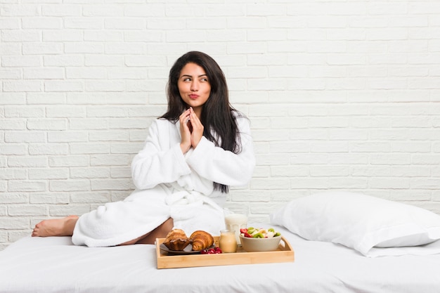 Giovane donna curvy che prende una colazione sul letto che compone piano in mente, installante un'idea.