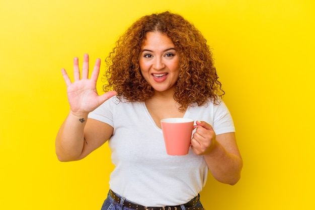 Giovane donna curvy latina che tiene una tazza isolata su fondo giallo sorridente allegro che mostra il numero cinque con le dita.