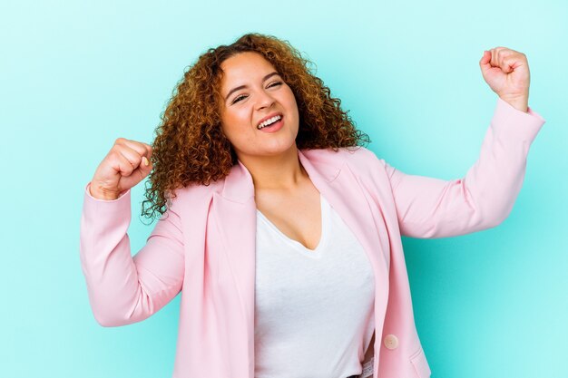 Giovane donna curvy latina isolata su priorità bassa blu che alza il pugno dopo una vittoria, concetto del vincitore.