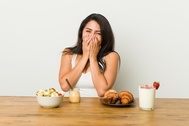 Giovane donna curvy prendendo una colazione ridendo di qualcosa, coprendo la bocca con le mani.