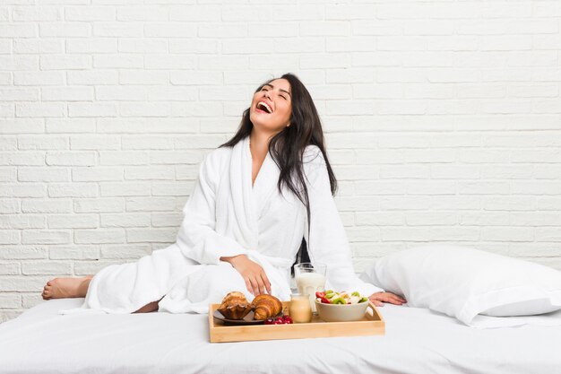 Giovane donna curvy prendendo una colazione sul letto rilassato e felice ridendo, collo allungato mostrando i denti.