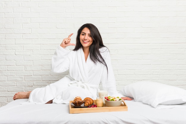 Giovane donna curvy prendendo una colazione sul letto tenendo qualcosa di piccolo con gli indici, sorridendo e fiducioso.