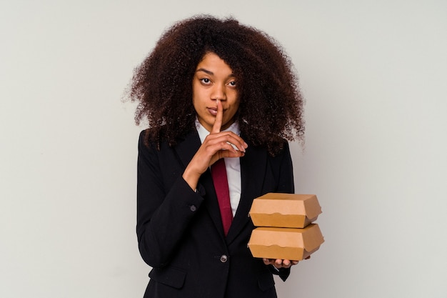 Giovane donna d'affari afroamericana che tiene un hamburger isolato su bianco mantenendo un segreto o chiedendo silenzio.