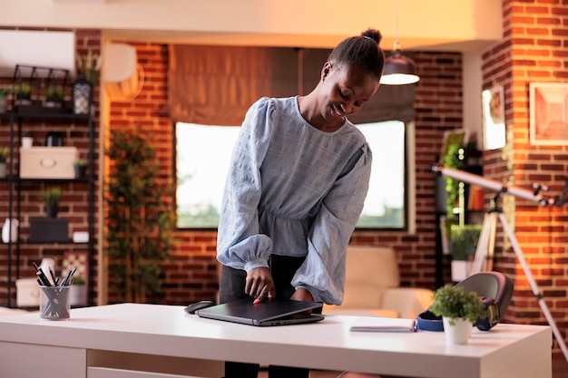 Giovane donna d'affari afroamericana sorridente che chiude il laptop e sta in piedi nella moderna sala di coworking. Libero professionista femminile che finisce la giornata lavorativa nel concetto di ufficio remoto dell'azienda