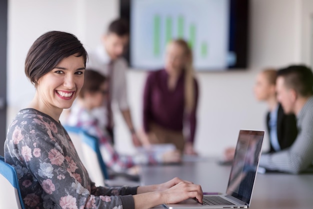 giovane donna d'affari all'interno dell'ufficio di avvio moderno che lavora su un computer portatile, squadra blured in riunione, gruppo di persone sullo sfondo