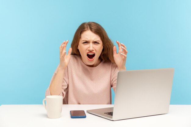 Giovane donna d'affari arrabbiata irritata in abiti casual che rugge e urla con odio espressione aggressiva furiosa seduta sul posto di lavoro home office indoor studio shot isolato su sfondo blu