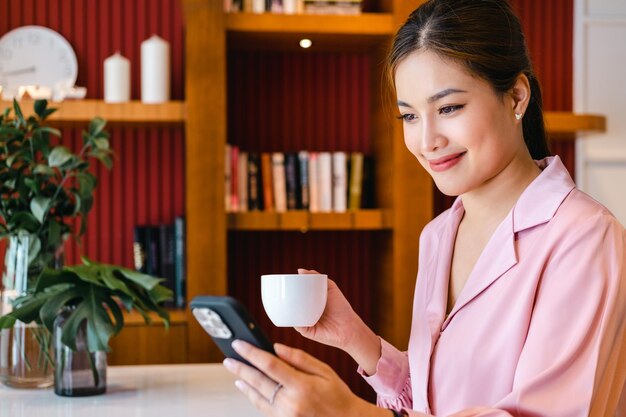 Giovane donna d'affari asiatica che beve caffè usando lo smartphone nell'ufficio di casa