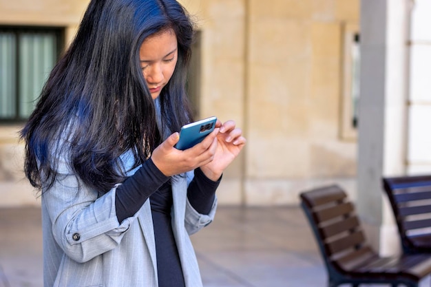 Giovane donna d'affari asiatica che indossa un abito in piedi sulla strada urbana utilizzando applicazioni sul cellulare, r