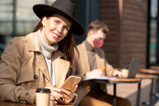 Giovane donna d'affari bruna elegante con cappello, maglione e trench che utilizza smartphone e ti guarda contro il networking dell'uomo