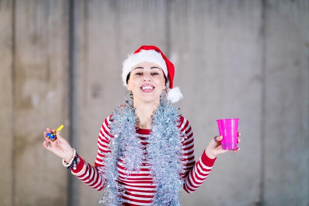 giovane donna d'affari casual felice che indossa un cappello rosso e soffia un fischio di festa mentre balla durante la festa di capodanno davanti al muro di cemento