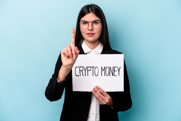 Giovane donna d'affari caucasica in possesso di un cartello di denaro crittografico isolato su sfondo blu che mostra il numero uno con il dito