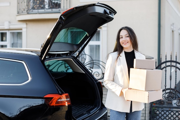 Giovane donna d'affari che preleva i pacchi da un bagagliaio di un'auto che torna a casa in auto Concetto di acquisto di merci online e di consegna a casa