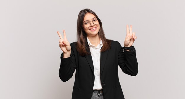 Giovane donna d'affari che sorride e sembra felice, amichevole e soddisfatta, gesticolando vittoria o pace con entrambe le mani