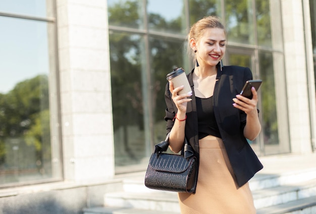 Giovane donna d'affari che utilizza smartphone e beve caffè mentre è in movimento all'aperto