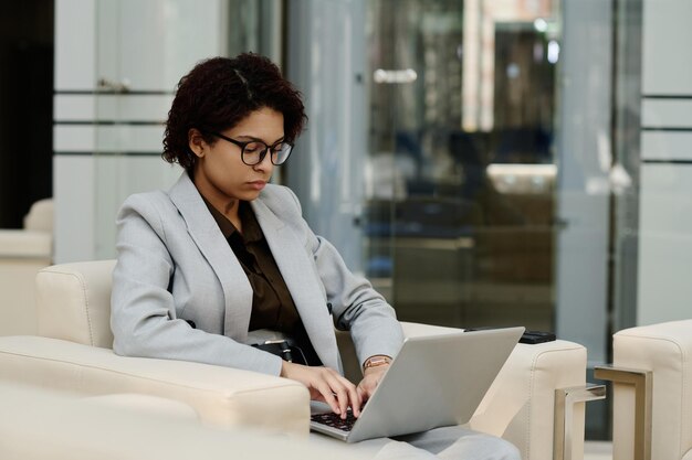 Giovane donna d'affari con gli occhiali che si concentra sul suo lavoro online su un computer portatile seduto su una poltrona