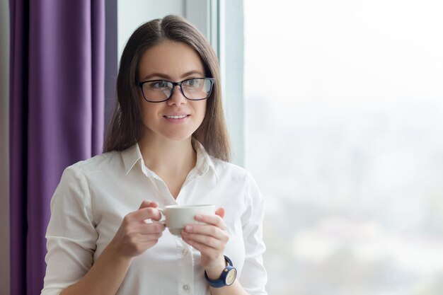 Giovane donna d'affari con una tazza di caffè vicino alla finestra in ufficio, città nella finestra per la stagione autunno inverno. Pausa caffè, insegnante donna positiva, psicologa, consulente, segretaria