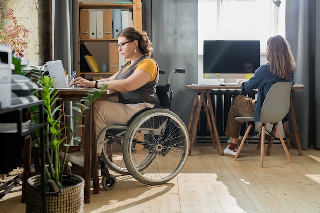 Giovane donna d'affari disabile in abbigliamento casual seduto su una sedia a rotelle davanti al laptop e alla rete mentre il suo collega lavora alla finestra