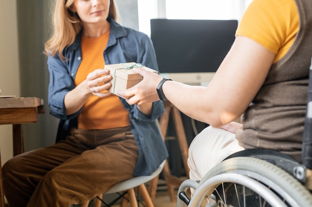 Giovane donna d'affari disabile in sedia a rotelle che passa una scatola regalo di cartone imballata con un regalo di Natale o di compleanno per la sua bella collega