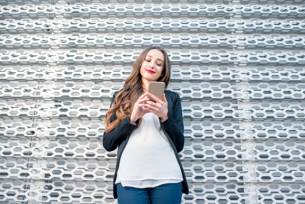 Giovane donna d'affari elegante nel vestito in piedi all'aperto con il telefono vicino al moderno muro di metallo