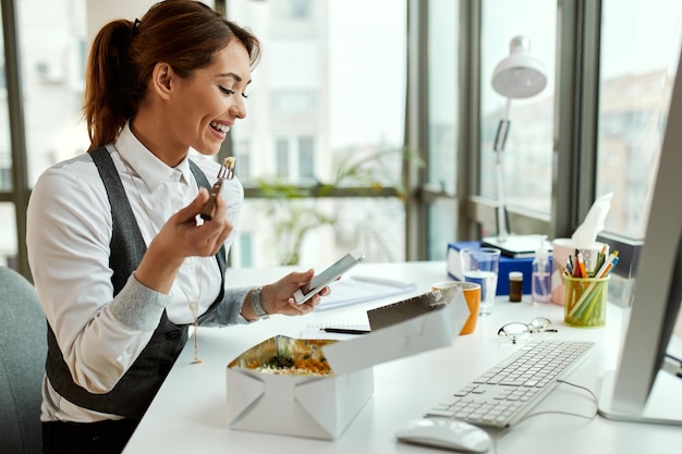 Giovane donna d'affari felice con pausa pranzo e messaggi di testo sul cellulare in ufficio