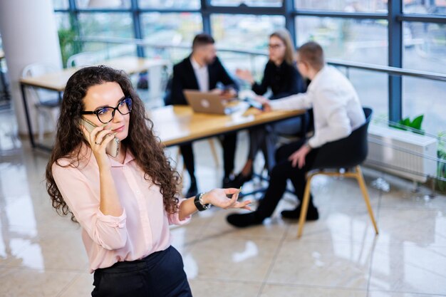 Giovane donna d'affari felice e intelligente, con abiti e occhiali eleganti, lavora in ufficio, parla con un cliente su uno smartphone, gesti, sorrisi. I dipendenti stanno lavorando in background. Messa a fuoco selettiva