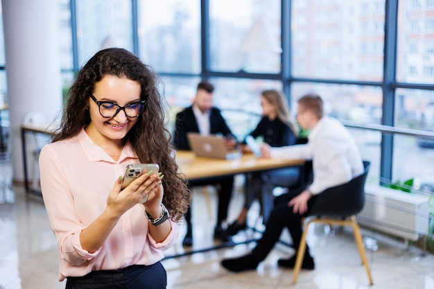 Giovane donna d'affari felice e intelligente, con abiti e occhiali eleganti, lavora in ufficio, parla con un cliente su uno smartphone, gesti, sorrisi. I dipendenti stanno lavorando in background. Messa a fuoco selettiva