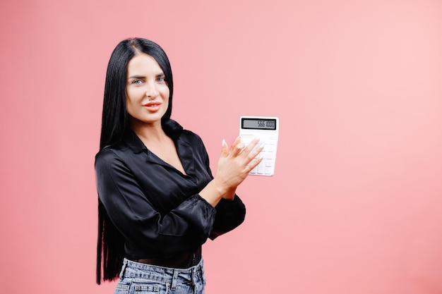 Giovane donna d'affari in camicia nera in posa isolata su sfondo rosa muro in studio Concetto di affari ricchezza carriera successo Mock up copia spazio La ragazza tiene in mano la calcolatrice