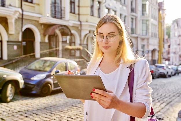 Giovane donna d'affari in città con tavoletta digitale. Bella donna sorridente in giacca bianca con occhiali guardando il monitor tablet, sfondo in stile urbano