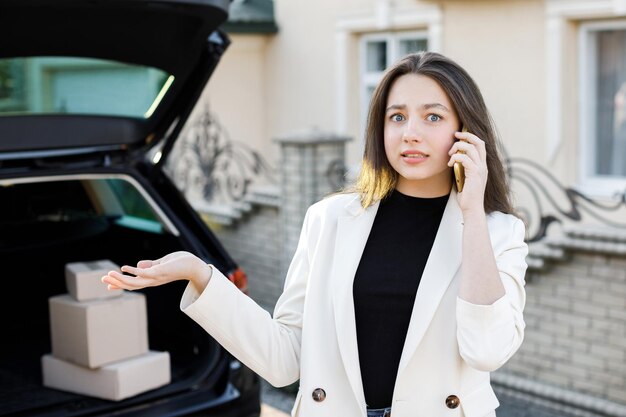 Giovane donna d'affari in piedi in macchina con pacchi che tornano a casa in auto La ragazza è in piedi e parla al cellulare vicino all'auto Concetto di acquisto di merci online e consegna a casa