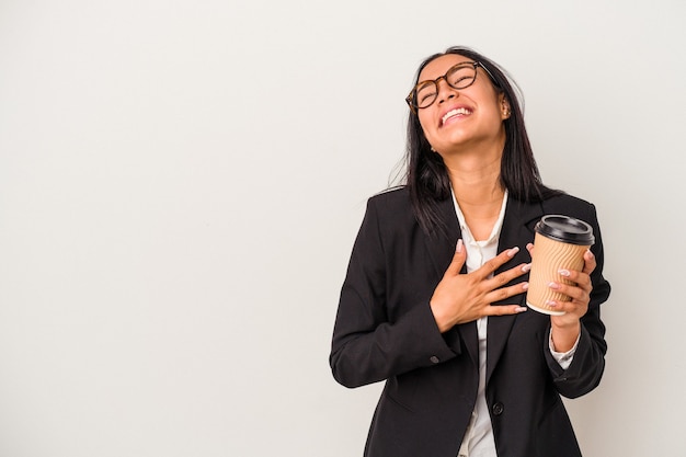 Giovane donna d'affari latina che tiene un caffè da asporto isolato su sfondo bianco ride forte tenendo la mano sul petto.