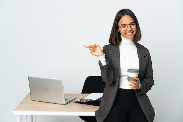 Giovane donna d'affari latino che lavora in un ufficio isolato sul muro bianco che punta il dito ai laterali e felice