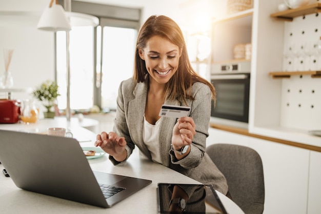 Giovane donna d'affari multi-tasking che utilizza il laptop e paga le bollette con la carta di credito a casa mentre si prepara per andare al lavoro.