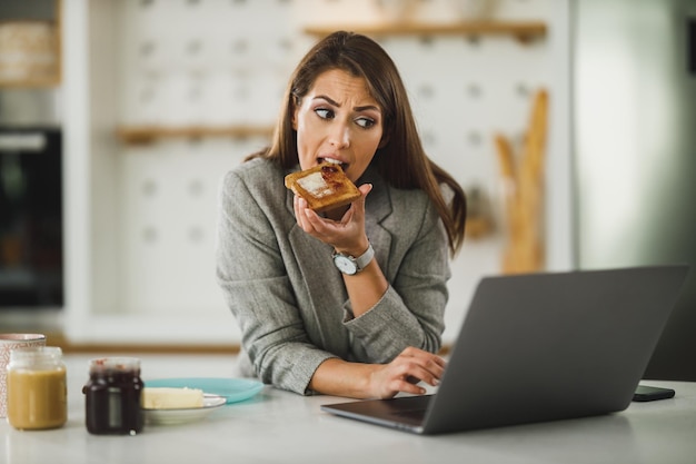 Giovane donna d'affari multitasking che fa colazione e usa il laptop mentre si prepara per andare al lavoro.