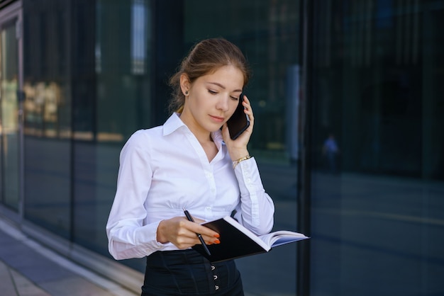 Giovane donna d'affari parlando al telefono e scrivendo su un taccuino. In una camicia bianca in piedi nell'edificio degli uffici