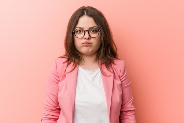 Giovane donna d'affari plus size caucasica soffia le guance, ha un'espressione stanca. Concetto di espressione facciale.