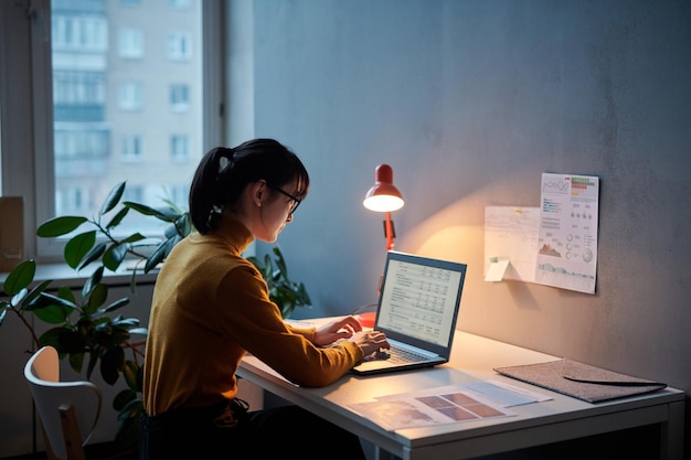 Giovane donna d'affari seduta al suo posto di lavoro e che lavora con il progetto online utilizzando il laptop che lavora fino a tardi in ufficio
