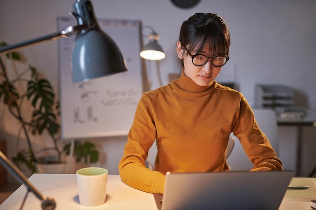 Giovane donna d'affari seduta al suo posto di lavoro e digitando sul laptop che lavora in ufficio fino a tarda sera durante la scadenza