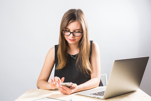 Giovane donna d'affari seduta in ufficio a tavola e utilizza lo smartphone. Sulla scrivania c'è un laptop. Donna che analizza i dati. Studenti che imparano online.