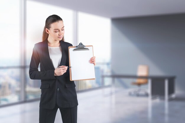 Giovane donna d'affari seria che indossa un abito che mostra ai suoi appunti con la penna in piedi in ufficio sfocato. Concetto di commercializzazione. Immagine tonica mock up