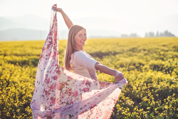 Giovane donna dai capelli lunghi che balla nel campo di fiori gialli, sciarpa che tiene in movimento dal vento, sfondo della retroilluminazione del sole pomeridiano