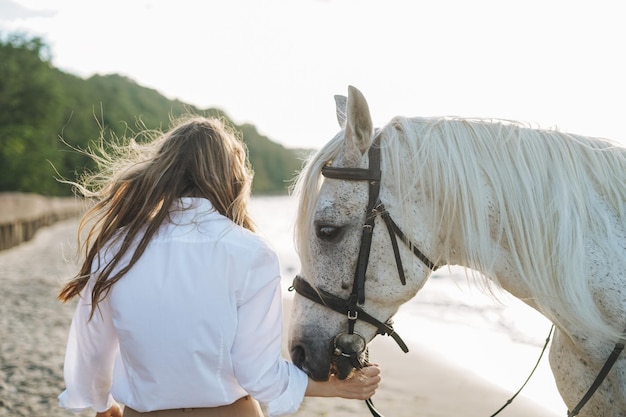 Giovane donna dai capelli lunghi in camicia bianca con cavallo bianco su sfondo marino