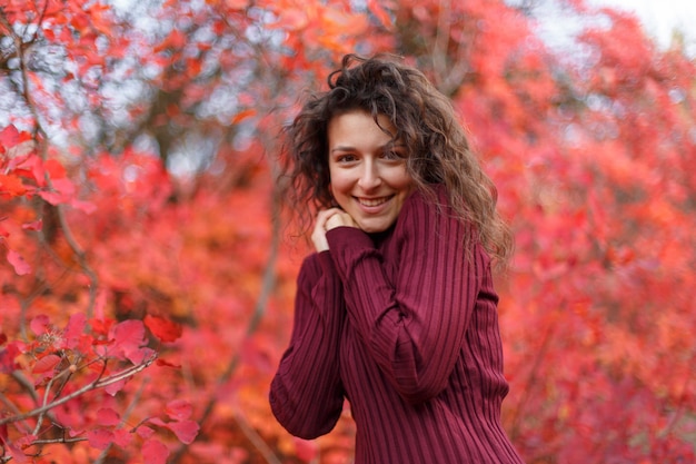 Giovane donna dai capelli neri in sweather rosso che riscalda le sue mani soffiando soggiorno in cespugli autumnn rossi.