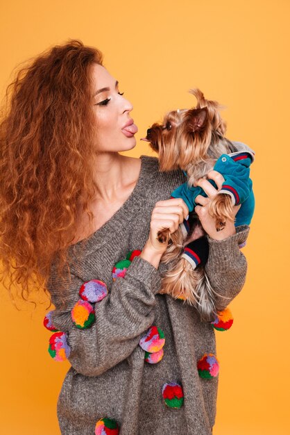 Giovane donna dai capelli rossi che mostra la lingua allo yorkshire terrier isolato sulla parete arancione