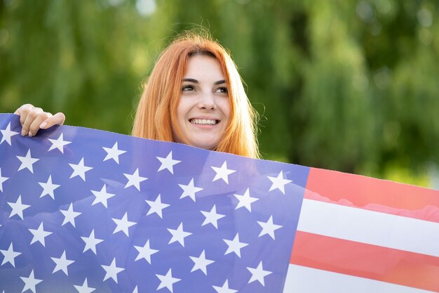 Giovane donna dai capelli rossi che tiene bandiera nazionale USA in piedi all'aperto nel parco estivo. Ragazza positiva che celebra il giorno dell'indipendenza degli Stati Uniti. Giornata internazionale del concetto di democrazia.