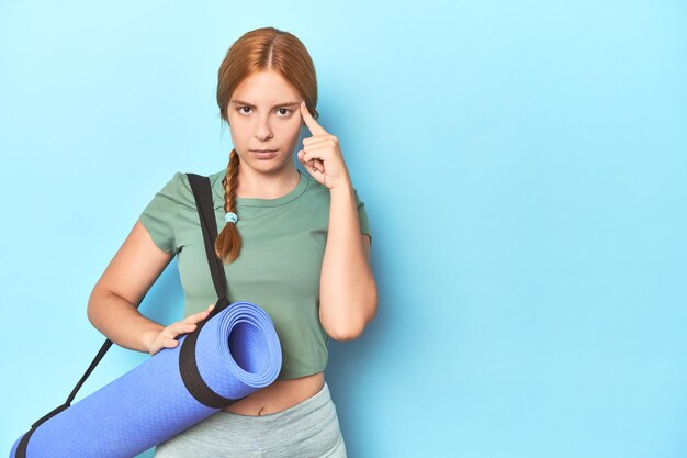 Giovane donna dai capelli rossi che tiene tappetino yoga in studio che punta il tempio con il pensiero del dito concentrato