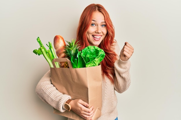 Giovane donna dai capelli rossi che tiene una borsa di carta con pane e alimentari che urla orgogliosa che celebra la vittoria e il successo molto eccitata con le braccia alzate