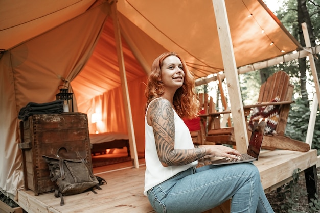 Giovane donna dai capelli rossi con un tatuaggio che usa il portatile vicino alla tenda glamping. Vacanza stile di vita in campeggio. Viaggio a basso budget, viaggio di vacanza nella foresta. Tecnologia di comunicazione delle informazioni di connessione Wi-Fi.