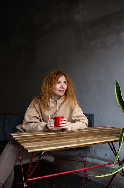 Giovane donna dai capelli rossi da sola che beve un drink in un caffè con terrazza in una giornata piovosa